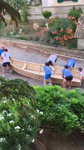 Seven young men, most wearing blue athletic shirts with white numbers, are carrying a large, light-colored boat. They are moving it up a sloped paved surface, possibly a street. The boat appears to be constructed of wood or a similar material and is very long. The men are positioned along the sides and underneath the boat, lifting and carrying it together. Some are running, while others are walking briskly. The environment includes green foliage, a building with a green shutter, a car parked in the background, and a potted plant. The overall scene suggests a collective effort to transport the boat.