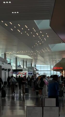 The video shows a busy airport concourse.  Many people with luggage are walking or standing, some looking at signs or information boards. There are shops and gate signs visible. The lighting is bright, with a patterned arrangement of lights across the ceiling. The overall impression is a crowded and active airport terminal.