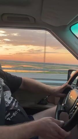 A first-person perspective video shows a person driving a car on a highway during sunset. The view outside the window is of a wide expanse of water with grassy islands, reflecting the vibrant orange and pink hues of the sky. The driver's hands are on the steering wheel. A black t-shirt with a graphic print is visible on the driver's arm. A black radio device is clipped to the inside of the car door. The sky is filled with dramatic clouds, painted with sunset colors.