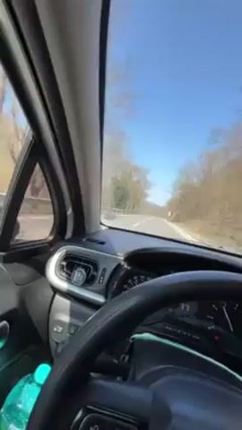 The video shows the interior of a car, specifically the driver's side, as it navigates a winding road on a sunny day. The car's dashboard, steering wheel, and a water bottle in the passenger seat are visible. Through the windshield, a clear blue sky and a road lined with trees are visible. The car is moving, and the scenery outside is shifting. The road has a guardrail on one side, and the surrounding terrain appears to be a hill or mountain covered with trees. Music is playing in the background.