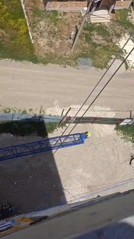 An aerial view shows a construction site with a blue crane. A person wearing a yellow hard hat and a green shirt is visible near the base of the crane. The crane appears to be in the process of being assembled or disassembled, with its arm extended. Various construction materials, including concrete blocks and metal pipes, are scattered around the site. A dirt road runs alongside the construction area, and a few buildings and vehicles are visible in the background. The ground is mostly dirt and gravel, with patches of dry grass and weeds.