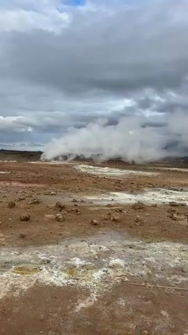 The video captures a barren, rocky, and muddy geothermal landscape in Reykjahlíð, Iceland, under an overcast sky. Large plumes of white steam billow from multiple vents across the scene, rising into the gray, cloudy sky. The ground is a mixture of reddish-brown earth and lighter, mineral-stained patches, indicative of the area's geothermal activity. Discolored pools and streams, likely heated by the vents, are visible throughout the terrain.

In the background, mountains, some with visible snow caps, are partially obscured by the steam and clouds. At the 0:02 mark, two small figures can be seen walking on a dirt path in the distance, heading towards the steam vents. A single person is also observed at the 0:03 mark, walking on the reddish-brown ground closer to a prominent steam vent. The time of day is daytime, despite the heavy cloud cover. The scene highlights the raw, powerful, and otherworldly natural phenomena characteristic of volcanic regions. No text is visible in the media.