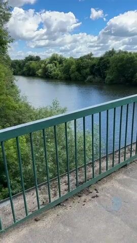 A stationary video shows a view from a bridge overlooking a wide, calm river. Green metal railings with vertical bars run across the foreground. The railings have some graffiti on them, including the numbers 2025 and 2026. Beyond the railings, the river stretches into the distance, flanked by lush green trees and foliage on both banks. The sky above is blue with fluffy white clouds. The ground in the immediate foreground of the bridge appears to be concrete and has some debris on it.