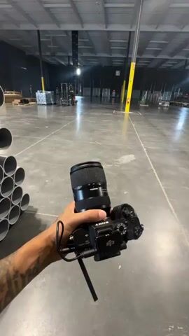This media file captures a person holding a Sony camera with a Tamron lens, filming within a large, industrial-style building. The scene appears to be a warehouse or a similar commercial space, characterized by its concrete floor, high ceilings, and exposed infrastructure like lighting rigs and metal beams.

In the foreground, a stack of large, gray pipes is visible to the left, indicating storage or construction materials. The camera operator, whose arm and tattooed forearm are in view, is actively filming.

The background shows several individuals, presumably workers or film crew members, engaged in various activities. One person in a red shirt is walking, and another in a blue shirt is standing near some large boxes. The overall atmosphere suggests an ongoing project or production, possibly related to real estate, construction, or event setup. The lighting appears to be a mix of natural light coming from large windows and artificial lighting from the ceiling fixtures, suggesting it could be daytime. There is no clear indication of weather conditions.

One of the boxes in the background has visible text that reads "DECORATIVE". The location is identified as El Paso, USA. The footage seems to be in motion, suggesting the camera operator is walking or moving around the space to capture different angles and areas. The presence of equipment and people working implies a busy, functional environment.