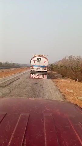 The video, taken from the perspective of a moving vehicle, shows a white tanker truck ahead on a cracked asphalt road. The tanker has an oval shape and is labeled "HIGHLY INFLAMMABLE" with the number 57 and the word "STOP" on either side of the number. The word "MASHAALLA" is written in large red letters on the mud flaps. A white van passes on the left and another red tanker truck appears in the distance behind it. The surrounding environment consists of dry, hazy scenery with sparse vegetation on the sides of the road. The sky is overcast.