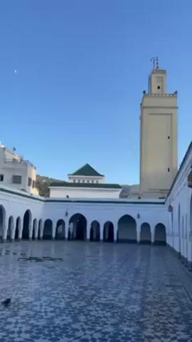 The video shows a courtyard with white arched walls and a blue and white tiled floor. In the background is a square white minaret, a green-roofed building, and a mountain range. The sky is blue. The camera slowly pans across the courtyard. There are people walking in the arched walkways. Hanging lamps are visible along the walls.