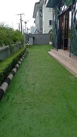 A short video clip shows an outdoor area with artificial grass. The area is bordered on the left by a grey concrete wall and a neatly trimmed hedge with a black and white striped border. Beyond the hedge are tall palm-like plants. A modern building with multiple stories is visible in the background, as well as power lines and streetlights. On the right side of the artificial grass is a building with a unique facade featuring green diamond-shaped patterns and large glass doors. Wooden steps lead up to this entrance. Several large white planters are placed around the area. The overall impression is that of a well-maintained, contemporary outdoor space.