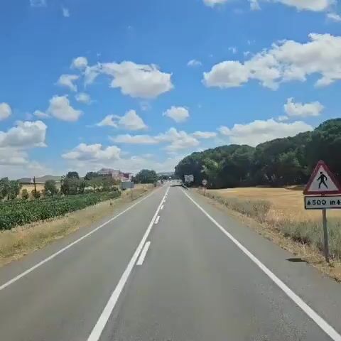 The video shows a view from a moving vehicle on a paved road. The road has white lines marking the lanes and a dashed white line in the center. On the left side of the road, there are vineyards and a building in the distance. On the right side, there is dry grass and trees. In the background, there is a large hill of earth or waste material, with a white structure on top that resembles a bird. There are also several road signs, including speed limit signs and directional signs. A white truck and a smaller white car are visible on the road ahead, approaching the viewer. The sky is blue with scattered white clouds. The overall scene appears to be a rural or semi-rural landscape on a sunny day.
