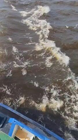 A video from the perspective of someone on a moving boat shows a wake trailing behind. The water is dark and choppy, with white foam churning in the wake. The edge of the boat is visible in the foreground, with blue and green surfaces. The overall impression is of a boat traveling at a moderate speed through rough water.