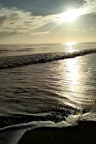 The video displays a tranquil seascape under a bright, somewhat hazy sun. The sunlight reflects prominently on the water's surface, creating a shimmering effect. Gentle waves roll in and break on the sandy beach in the foreground. The sky is partially covered with clouds, which adds depth and texture to the horizon. There are no visible people or objects, the focus is strictly on the natural scenery.