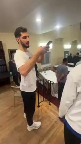 A man in a white t-shirt and black pants holds a phone and shows it to another man wearing a white sweatshirt. They are standing in a room with light hardwood flooring, and a maroon carpet is visible in the background. Several people are walking around in the background, and tables and chairs are set up in the room. There is also a framed piece of art on the wall. The room is well-lit. The man with the phone appears to be smiling.