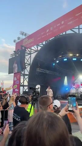 This video captures an outdoor music concert on a stage set up for a "Day of Youth" festival, likely taking place in Novosibirsk, Russia, given the Cyrillic text on the banner. The time of day appears to be late afternoon or early evening, with a clear sky and soft, warm lighting from the setting sun illuminating the clouds.

The primary subjects are a large crowd of young people gathered to watch a performer on stage. The performer, dressed in light-colored clothing, is visible on the main stage and also on a large LED screen above the stage. The stage itself is elaborate, featuring a curved roof structure, lighting rigs with blue and pink lights, and large speaker arrays. A videographer is actively filming the performance with a professional camera and tripod positioned in front of the crowd. Many audience members are also recording the event on their mobile phones.

The atmosphere seems energetic and celebratory, with people actively engaged in the performance, many holding their phones aloft. The visible text on a large red banner above the stage reads "ДЕНЬ МОЛОДЁЖИ" which translates to "Day of Youth," confirming the nature of the event. Other smaller logos and text, though partially obscured, suggest sponsorships or organizational affiliations. The overall scene depicts a lively and engaging youth festival in an urban outdoor setting.