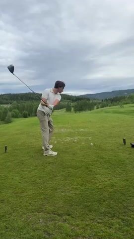 A man is playing golf on a green, grassy course. He is wearing a white polo shirt, beige pants, and white sneakers. He swings a golf club, hitting a white golf ball. The sky is cloudy and overcast. In the background, there are green trees and hills. The man watches the ball fly after hitting it, then sets his club down.