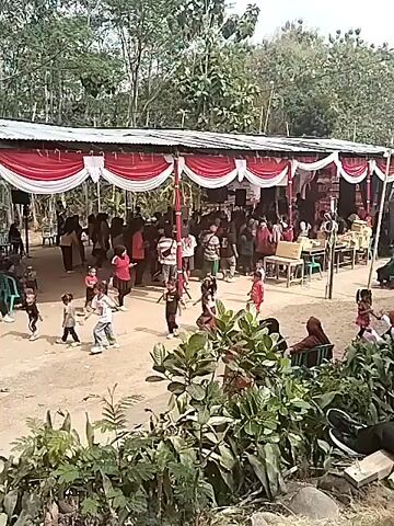 The video shows an outdoor gathering, likely a celebration or event, with a large tent set up. The tent is decorated with red and white draped fabric. Many people of various ages are present, some standing, some sitting, and some dancing. In the foreground, several children are visible, some dancing and playing. In the middle ground, a group of adults are gathered under the tent, some appearing to be watching or participating in an activity. There are tables and chairs set up under the tent, suggesting food or seating arrangements. A speaker system is also visible, indicating music or announcements are being made. The environment appears to be rural, with trees and foliage surrounding the area. The ground is dirt.