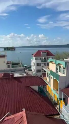 The video shows a view of a town with a body of water in the background under a sky with scattered clouds. The foreground is filled with colorful buildings with red roofs. The buildings are a mix of sizes and architectural styles. Some have white walls and red roofs, while others are painted in yellow and turquoise. Clothes can be seen hanging out to dry on the roofs of some buildings. The water in the background is calm, with forested land visible on the opposite shore.