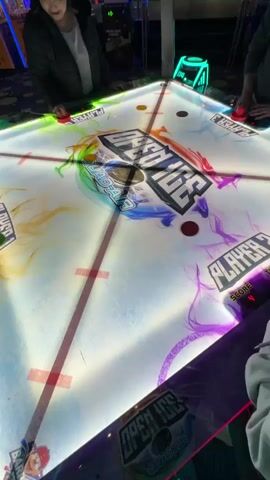 This video captures four individuals playing a vibrant game of four-player air hockey on an "Open Ice Quad Air" table within an arcade setting in Schaumburg, United States of America. Each player holds a red mallet, actively striking multiple colored pucks – including yellow, green, and red – across the illuminated table.

The air hockey table is the focal point, featuring bright LED lighting that casts neon green, red, and purple hues, enhancing the dynamic gameplay. The table's surface is adorned with colorful, swirling graphics and prominent text displaying "OPEN ICE QUAD AIR" in the center. Player designations "PLAYER 1" and "PLAYER 2" are visible, along with digital score displays indicating "SCORE" and "TIME," with "PLAYER 2" showing a score of "4."

The players are engaged in intense, fast-paced action, moving their mallets quickly to hit the pucks. The background offers glimpses of other arcade machines, confirming the entertainment venue setting. The lighting is artificial, typical of an indoor amusement area. The sounds suggest an energetic environment with game effects and casual chatter from the participants. The atmosphere appears lively and competitive as the game unfolds.