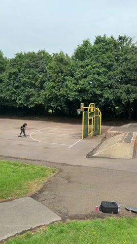 A young adult male is captured in an outdoor setting on a cloudy day, engaged in scooter riding. The individual, dressed in a black t-shirt and loose-fitting pants, rides a black scooter toward a concrete ramp. He rides up the ramp and then lands back onto the flat ground. The scene is located in a park-like area, featuring a basketball court with a distinctive yellow and green backboard and hoop. Adjacent to the ramp is a smaller concrete structure. A green trash can is visible near the treeline. Personal items, including a black backpack, a water bottle, and a skateboard, are placed on the grass near the edge of the paved area.