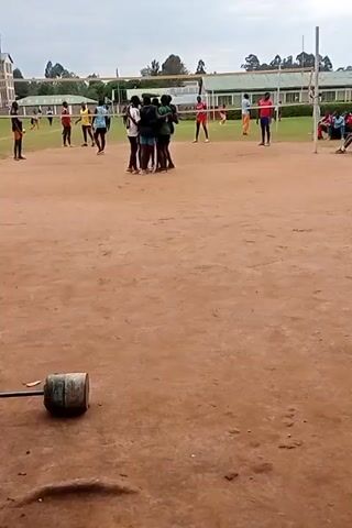 The video shows a volleyball game being played on a dirt court. There are several players visible, wearing different colored shirts and shorts. They are divided into two teams, one wearing green shirts and the other wearing a mix of colors, including yellow, blue, and white. In the background, there is a building and some trees. In the foreground, there is a metal object lying on the ground. The players are actively engaged in the game, hitting the ball over the net and moving around the court. Some of the players are grouped together, possibly celebrating a point. The overall environment suggests an outdoor sports activity taking place on a somewhat cloudy day.