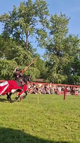 Two individuals on horseback, dressed in medieval armor and red and white caparisons, charge at each other with lances in a jousting reenactment. They are positioned on a grassy field with a wooden fence and spectators in the background. Trees and a clear blue sky are visible.