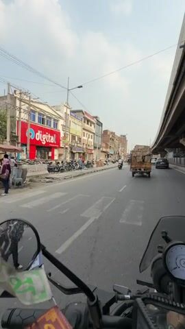 This video captures a first-person perspective of a motorcycle ride through a bustling street in Panipat, India. The rider, wearing a red and black helmet and sunglasses, is the primary subject in the foreground. The scene is a road with multiple lanes, bordered by buildings on the left and an elevated highway structure on the right.

The activity is a motorcycle journey. The rider is moving forward, indicated by the road ahead and the passing scenery. The environment appears to be urban, with shops and businesses lining the street. On the left, we see a row of shops with signage, including a prominent red sign with white lettering that appears to say "Odigal". Other signs are in Hindi. A variety of motorcycles are parked along the sidewalk.

On the right side of the road, traffic moves under an elevated highway. A yellow truck carrying goods is visible ahead. Other vehicles, including cars and motorcycles, are also present on the road. The sky is partly cloudy, suggesting a daytime setting, likely morning or afternoon. The overall atmosphere seems lively and characteristic of a busy Indian city street.

The video showcases the dynamic street life of Panipat, with a mix of commercial activity and vehicular traffic. The focus on the rider and their journey provides a sense of immersion in the experience. The text visible on the rider's shirt appears to be in Cyrillic script, reading "СВОБОДНЫЙ".
