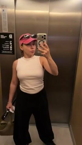 This media file is a short, silent video clip of a young woman taking a mirror selfie in an elevator.

**Subjects:** The primary subject is a young woman with blonde hair pulled back in a braid. She is wearing a white sleeveless top, black loose-fitting pants, a pink baseball cap, and black sunglasses perched on top of the cap. She is holding a dark-colored object, possibly a water bottle, and a tan handbag in her left hand. She is also holding a smartphone in her right hand, which she is using to take the selfie.

**Scene/Setting:** The setting is the interior of a modern elevator with brushed metal walls. The elevator doors are visible, and the button panel with illuminated numbers can be seen on the right-hand side. There is a sign with text on the left-hand wall.

**Activity/Event:** The woman is in the process of taking a selfie. Her body is angled slightly, and she is looking at her phone's screen. The elevator appears to be stationary at the moment the video is captured.

**Notable Details:** The lighting in the elevator is artificial and appears to be ambient, suggesting it could be any time of day. There are no strong indicators of weather or specific emotions, though her gaze towards the phone suggests a focus on capturing the image. The presence of the elevator suggests she is in a multi-story building. The location context provided, Žunovnica, Bosnia and Herzegovina, offers a geographical setting, but there are no visual cues within the frame to confirm this location.

**Visible Text:** On the left-hand wall of the elevator, there is a sign with text in a small font. The text is partially obscured and difficult to read clearly, but it appears to be a set of instructions or information typically found in an elevator. There is also a logo that is partially visible.