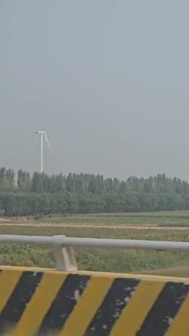 The video shows a landscape with a wind turbine in the distance, situated behind a line of trees. In the foreground, there is a barrier with yellow and black stripes. The sky is overcast. The video appears to be taken from a moving vehicle. The text overlays on the video indicate that the turbine's design is from Hebei, China, and it is not a repeated image.