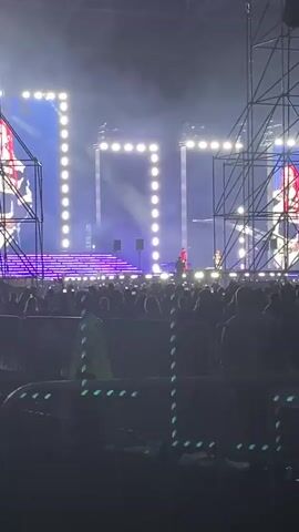 A wide shot captures a lively concert scene. In the background, a stage is illuminated with bright lights and a large screen displaying an animated image. Two figures, possibly performers, are visible on stage, one in red and another in black. A large crowd of people fills the foreground, many with their backs to the camera, suggesting they are facing the stage. The atmosphere appears energetic, with a sense of excitement emanating from the audience. The lighting is dramatic, with spotlights and stage effects creating a vibrant ambiance. Metal barricades separate the crowd from the stage area, and security personnel in yellow vests are positioned among the attendees. The overall impression is of a large-scale musical event.