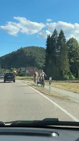 The video is shot from the perspective of someone in a vehicle, looking forward. A paved road is in the foreground, with a white line separating lanes. To the right of the road is a paved bike path. On the bike path, a man is riding a bicycle, pulling a small trailer. Next to him, also on the bike path, a child is riding a bicycle. On the grassy area to the right of the bike path, a scarecrow is standing. The scarecrow is wearing an orange shirt and has a straw hat. It is holding a long stick. In the background, there are houses and a wooded hill under a bright blue sky with scattered white clouds. A black car is visible in the distance on the road ahead.