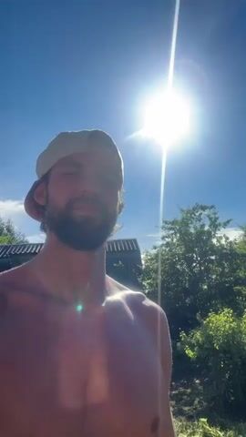 A shirtless man wearing a baseball cap is filmed outdoors. The video begins with a shot of the man looking up at the sun, which is bright and in the upper right portion of the frame. He is standing in front of a dilapidated building with a corrugated metal roof. In the next shot, the man turns his head and smiles at the camera. The background shows lush green foliage. The video then shows a close-up of the man's face as he squints slightly. The final shot is a medium shot of the man as he stands in front of the building again, with a wide smile. The building appears to be made of concrete blocks and has a wooden door. There are also some wooden planks on the ground in front of the building. The sun is still visible in the sky, casting a shadow on the ground.