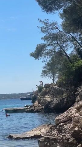 The video shows a coastal scene on a clear, sunny day with two people standing on a rocky cliff overlooking the ocean. The rocky cliff is covered with vegetation, including tall pine trees that partially frame the scene. The ocean water is a clear turquoise color, and there is a small boat anchored near the shore. In the distance, a larger boat can be seen on the horizon. One person jumps off the cliff into the water.