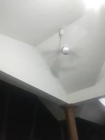 The video captures a white ceiling fan with three blades, suspended from a white ceiling by a rod. The fan is stationary. The ceiling is angled and painted white, with shadows cast by the fan and possibly the structure of the ceiling itself. In the lower right quadrant of the frame, a portion of a wooden pillar is visible. The background is dark, suggesting an indoor environment, possibly a room with dark walls or the absence of light in that area.