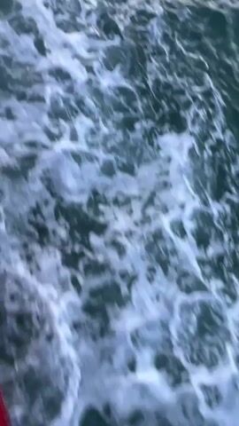This video shows a view from a boat moving through the ocean. The water churns into white foam in its wake, indicating movement. In the distance, a calm sea stretches towards a horizon lined with silhouetted hills. The sky above is filled with dramatic clouds, with streaks of orange and yellow suggesting either sunrise or sunset. The overall mood is tranquil and contemplative, evoking the feeling of a peaceful journey at sea. The time of day is likely either early morning or late evening. The location is identified as Thong Nian, Thailand. No people are visible in the frame.