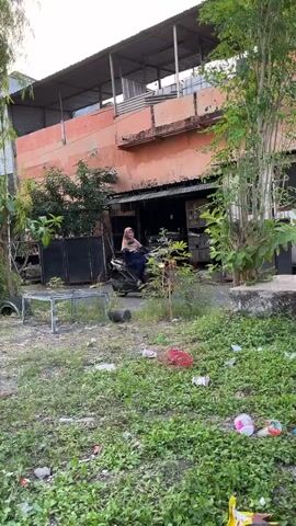 The video shows a street view with a building and some greenery. In the foreground, there is a patch of overgrown grass with some scattered trash, including plastic bottles and wrappers. A small metal cage or stand is visible to the left.

In the mid-ground, a person wearing a hijab is seated on a motorcycle, parked in front of a building. The building appears to be a multi-story structure with an orange-colored facade. There are open doorways or garage-like entrances on the ground floor. Some trees and bushes are present around the building.

The background shows more of the building and some vegetation. The overall scene suggests an outdoor environment. The lighting indicates it might be late afternoon or early evening, with soft sunlight. The location context of Kalisampurno, Indonesia, suggests a tropical or subtropical setting. No discernible text is visible in the frame. The mood appears neutral and observational, focusing on a simple street scene.
