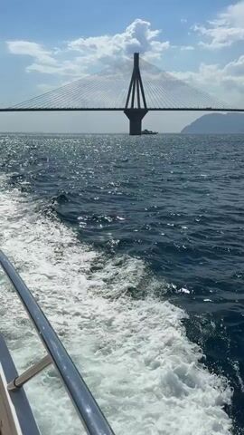This is a vertical, first-person perspective video taken from a boat on the water. The scene is a bright, sunny day with scattered white clouds against a blue sky. The water is a deep blue with white foamy waves trailing behind the boat, indicating movement. In the distance, a large cable-stayed bridge spans across the water, connecting landmasses on either side. The bridge is characterized by its tall, V-shaped pylons and numerous cables that support the roadway. A ship is visible in the distance, beneath the bridge. The overall mood is serene and picturesque, suggesting a leisurely boat trip on a beautiful day in a coastal area. The Municipal Unit of Rio, Greece, provides geographical context, implying the bridge is likely the Rio-Antirrio Bridge, a significant landmark in the region. There is no visible text in the video.