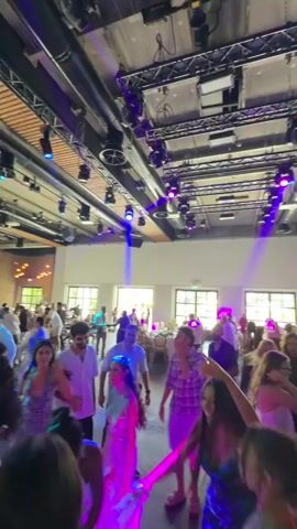 The video shows a lively indoor event with a large group of people dancing and socializing. The environment appears to be a spacious venue with a high ceiling, exposed beams, and industrial-style lighting. Multiple spotlights cast colorful beams of purple and blue light across the room, creating a festive atmosphere. People of various ages and ethnicities are dressed in casual and semi-formal attire, engaging in dancing and conversation. Some individuals are holding drinks. The overall mood is one of celebration and enjoyment, suggesting a party, wedding reception, or similar gathering.