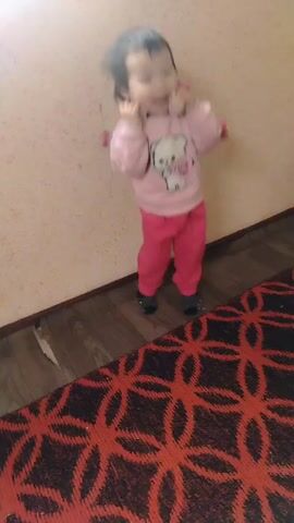 A young child with dark hair stands in a corner between a light-colored wall and a dark wooden floor. The child is wearing a pink sweater with a white teddy bear print and pink pants with dark socks. The child is holding a pink stuffed toy up to their ears as if it were a telephone receiver. There is a red and black patterned rug partially visible in the lower right corner of the frame. The child is the only person visible, and they appear to be talking into the toy. The wall has some slight damage near the base. The room is sparsely decorated and illuminated.