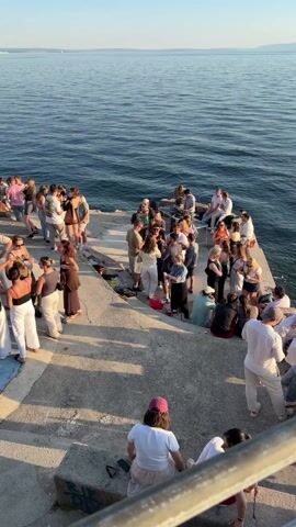 The video depicts a gathering of people on a concrete pier or platform adjacent to the ocean. Numerous individuals, mostly young adults, are seen standing, sitting, and socializing. They are dressed in casual, summer-appropriate attire. A DJ booth is set up on the pier, with two people seemingly operating the equipment. The ocean stretches out behind them, with distant land visible on the horizon. The camera pans to reveal the sun reflecting off the water. The weather is clear and sunny. The overall atmosphere suggests a relaxed, social gathering or party.