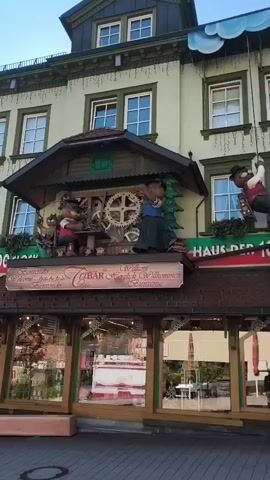 The video displays a large, ornate cuckoo clock on the facade of a building. The clock features animated figures, including two bears and a man in a red vest, who appear to be engaged in various activities. The bears are positioned at the clock's mechanism, with one seemingly operating gears. A man is depicted hanging from a rope suspended from a cloud above the clock. Below the clock, a sign reads "CUBAR". The building itself is multi-story with visible windows, and at street level, a storefront with large windows showcases merchandise. The overall setting suggests a commercial area, possibly a town square or a street known for its unique attractions. The lighting indicates it may be daytime, with a bright sky visible at the top of the frame.