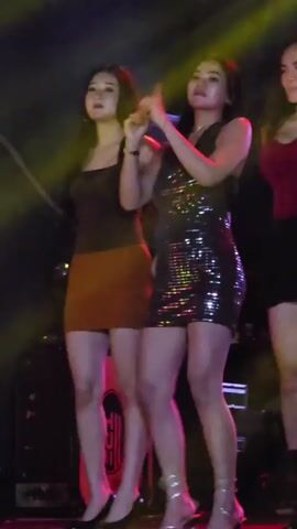 Three young women are standing on a stage, likely performing. The woman on the left wears a brown top and an orange skirt. The woman in the middle wears a black sequined dress. The woman on the right wears a red top and a black skirt. They are all looking forward or to the side, and the woman in the middle is gesturing with her hands. The stage is lit with colored lights, and there are some speakers or equipment visible in the background.