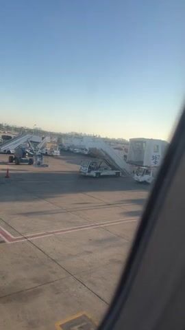 The video shows an airport tarmac on a clear day. Several mobile staircases are positioned next to parked aircraft, suggesting boarding or deplaning operations. The staircases are white and have visible numbers and text, with one clearly marked "367". Small service vehicles, likely tugs or baggage carts, are attached to some of the staircases. The ground is asphalt, marked with red and yellow lines. The sky is a clear blue, and sunlight casts shadows across the tarmac. A blurry edge of what appears to be an airplane window frame is visible in the foreground, indicating the video was filmed from inside an aircraft. No people are clearly visible on the tarmac.