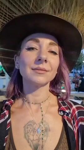 The video is a selfie-style recording of a woman wearing a hat and a plaid shirt. She has purple hair, a tattoo on her chest, and is wearing necklaces. In the background, there is a beach and the ocean, with some people sitting at tables in what appears to be an outdoor dining area. The lighting suggests it is late afternoon or early evening.