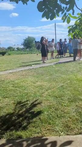 A group of people are gathered outdoors on a sunny day. Some are standing and talking, while others are seated. The environment appears to be a lawn with a building in the background. One person is taking a photo with their phone.