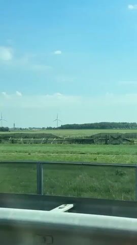 The video captures a view from inside a vehicle, looking out at a rural landscape under a bright blue sky with scattered clouds. In the foreground, a metallic railing is visible, partially obscuring the view. Beyond the railing, a grassy field stretches out, with some plants and vegetation. Further in the distance, there are more fields and trees. Two wind turbines are prominently featured, one on the left side of the frame and another on the right, with their blades visible. There are also some low structures and possibly fences in the middle ground. The overall impression is of a sunny day in an open, agricultural area.