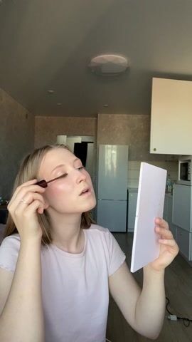A young woman with fair skin and blond hair is applying mascara to her upper eyelashes. She is holding a small, rectangular mirror in her left hand to aid in the process. She is wearing a light pink t-shirt and a simple ring on her left ring finger. The background reveals a neutral-toned room, seemingly an apartment. Behind her, there is a kitchenette with white cabinets, a refrigerator, and a microwave. The lighting is bright and appears to be natural daylight. The room is somewhat cluttered with a few items visible on a nearby surface.