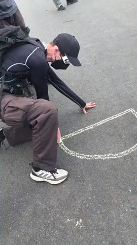 A man is drawing on asphalt with chalk. He is kneeling on his left knee and extending his right arm to draw. The man is wearing a black long-sleeve shirt, brown pants, and Adidas athletic shoes. He also has on a black baseball cap and a black face mask. A black backpack is strapped to his back. He is drawing a shape that could be a "D," or a "P," or potentially a peace sign. Parts of other people are visible in the background, including legs and shoes. The ground is asphalt.