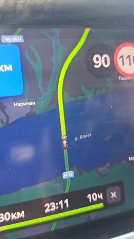 The video shows a car traveling across a bridge, indicated by the Volga river on the navigation screen. The navigation screen also shows a speed of 90 km/h within a 110 km/h limit. The exterior shot reveals a body of water and trees in the distance. The view shifts to the car's interior, capturing the rearview mirror, a portion of the dashboard, and the side window. A person wearing a black baseball cap is seen in the passenger seat.
