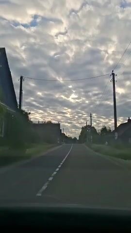 The video shows a first-person perspective of driving down a paved road. The road is surrounded by trees and grass. There are power lines running along the side of the road. The sky is cloudy with patches of sunlight breaking through. The time stamp indicates the video is playing at normal speed.