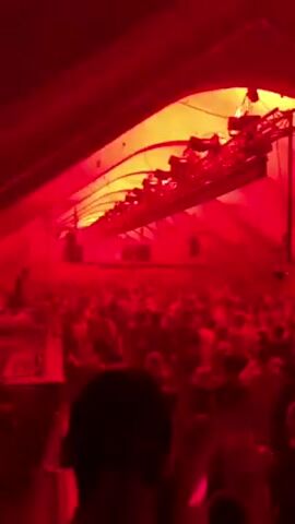 A crowd of people dances under flashing white lights in a large, dark, indoor space. There are multiple speakers and spotlights mounted on the ceiling. A red laser beam flashes above the crowd. The general atmosphere is energetic and festive.
