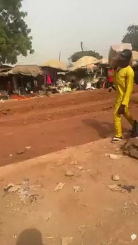 The video shows an outdoor marketplace in a developing area. The ground is unpaved and covered in reddish-brown dirt, rutted with tire tracks. A man in a yellow outfit and black cap carries a large cardboard box on his shoulder. He walks past the camera, revealing a view of market stalls with covered awnings. People mill around the stalls, some carrying shopping bags. In the distance, construction equipment, including a yellow bulldozer and a dump truck, are visible. A tall communications tower stands in the background. The weather appears hazy or dusty.