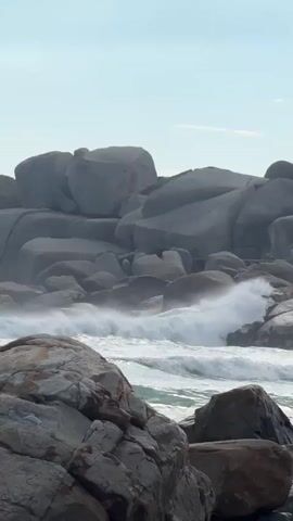 The video shows a rocky coastline with large gray boulders. Waves crash against the rocks, creating white foam. The sky is light blue with a few faint clouds.